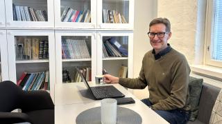 Henkilö istuu pöydän ääressä kahvikupin ja tietokoneen kanssa. Taustalla on valkoinen kirjahylly.